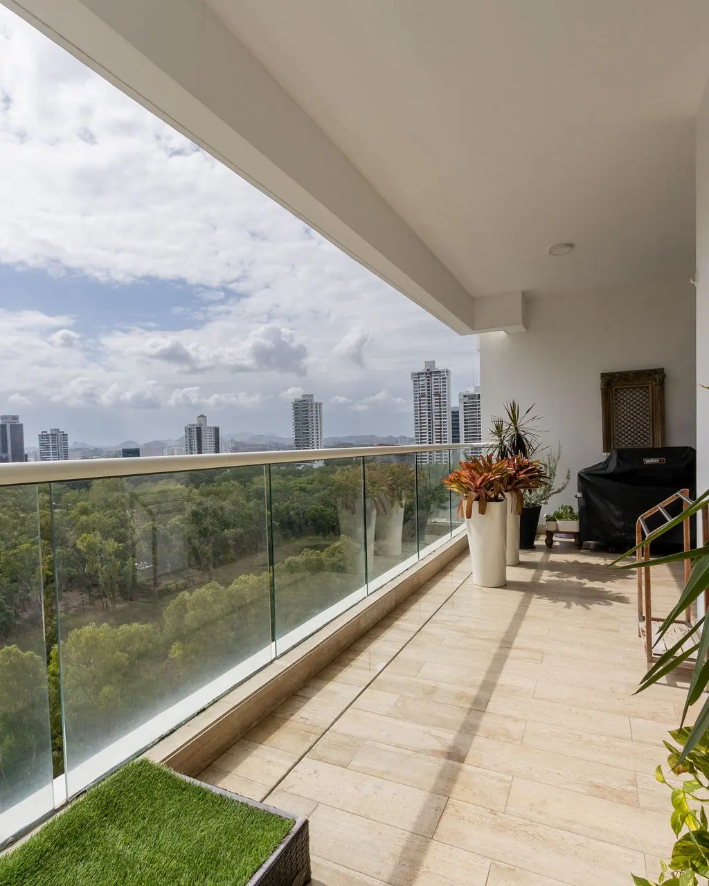 A bright, modern balcony with glass railings overlooks a green landscape and city skyline. Potted plants and outdoor furniture add to the inviting space.