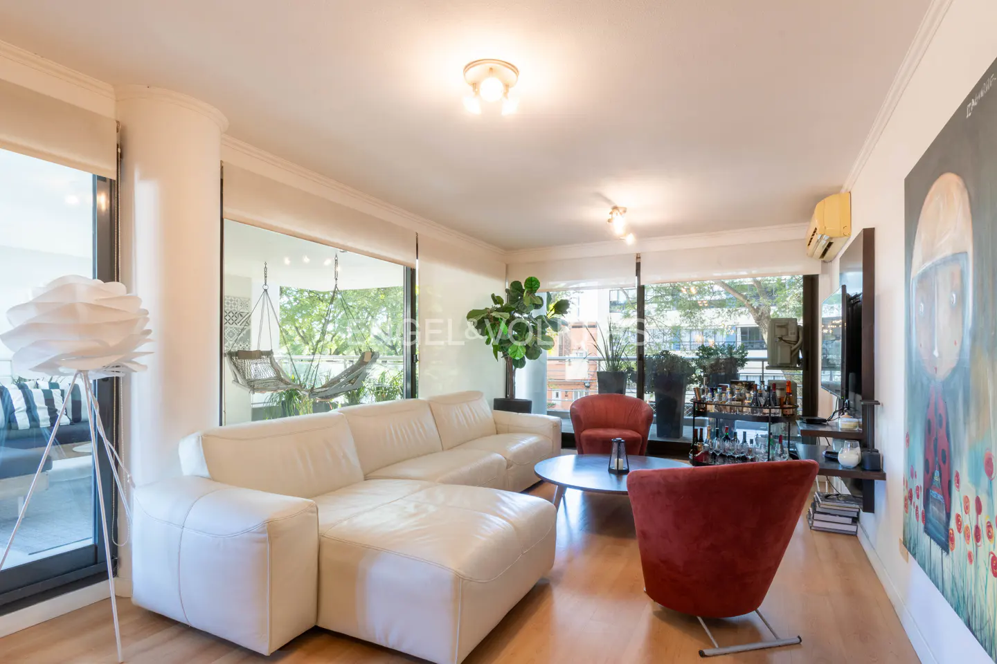 Bright living room with a white leather sectional sofa, red chairs, and a round coffee table. Large windows offer views of trees and a hammock.