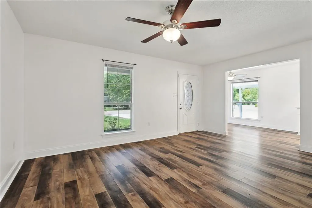 Bright, empty living room with wood floors, white walls, a ceiling fan, a window, and a white front door.