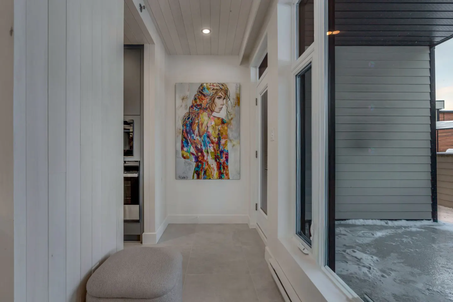 Hallway with white walls, tile floor, and a colorful painting of a woman. A window shows a snowy patio.