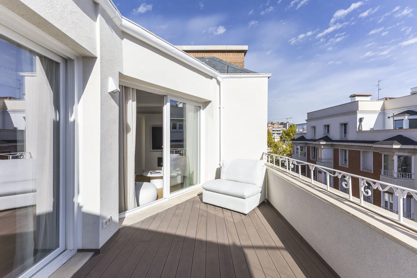 A bright, modern balcony with a white chair, wood-look flooring, and a white railing overlooking city buildings under a blue sky.