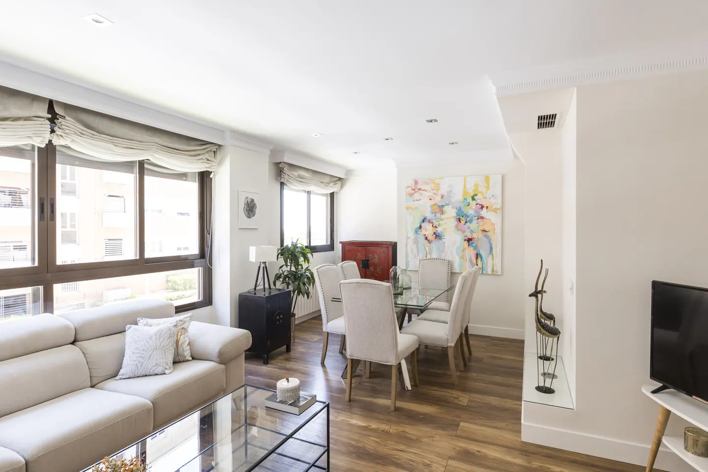 Bright living room with a beige sofa, glass coffee table, dining table with six chairs, and a colorful abstract painting.