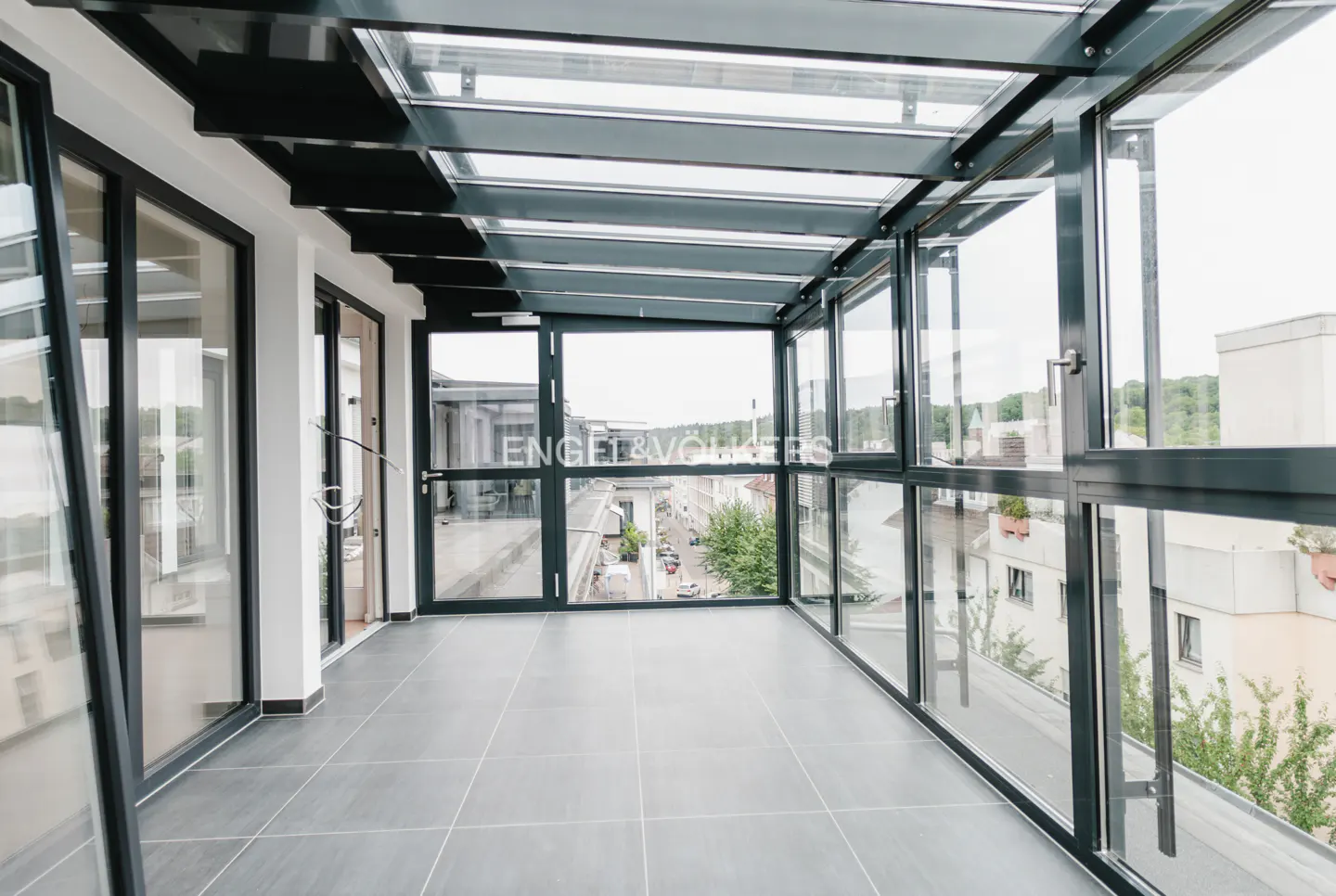 Modern enclosed balcony with gray tile flooring, black metal frames, and a glass roof overlooking a city street.