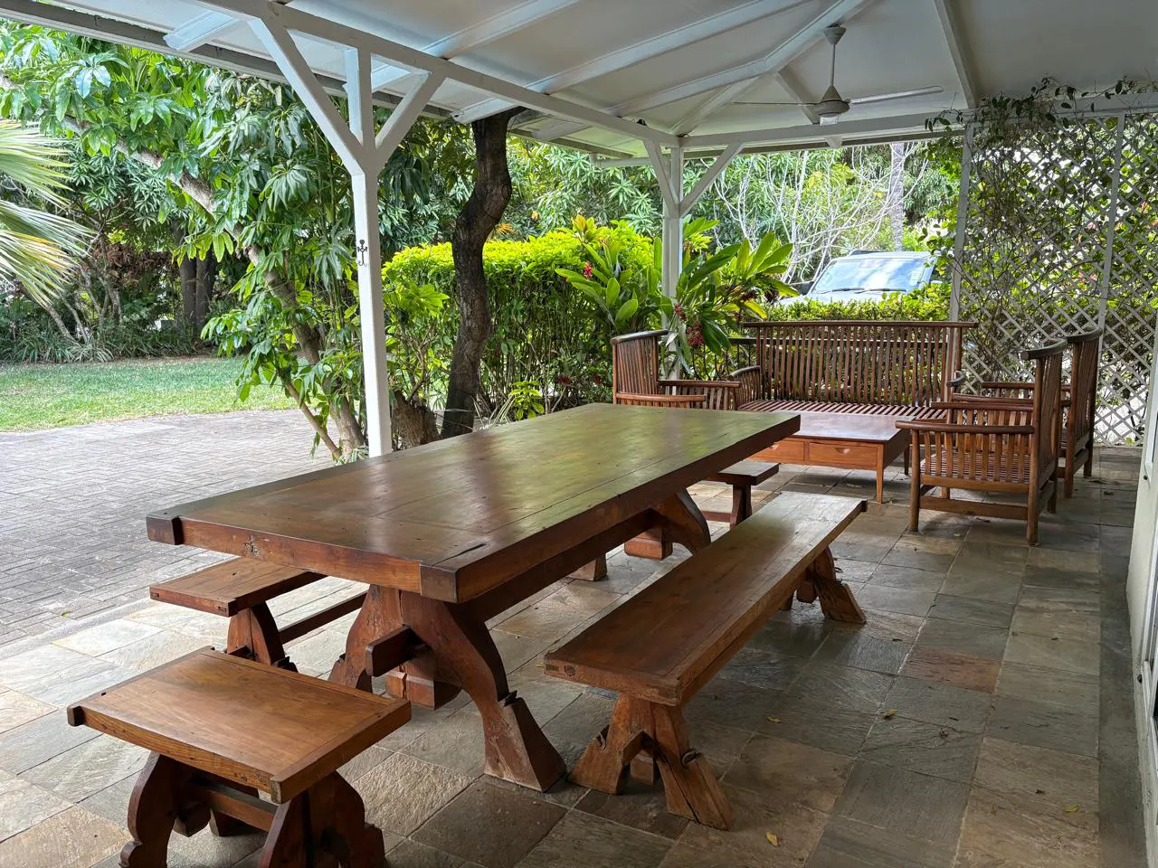 Covered patio with a large wooden dining table, benches, and chairs on a stone floor, overlooking a lush green garden.