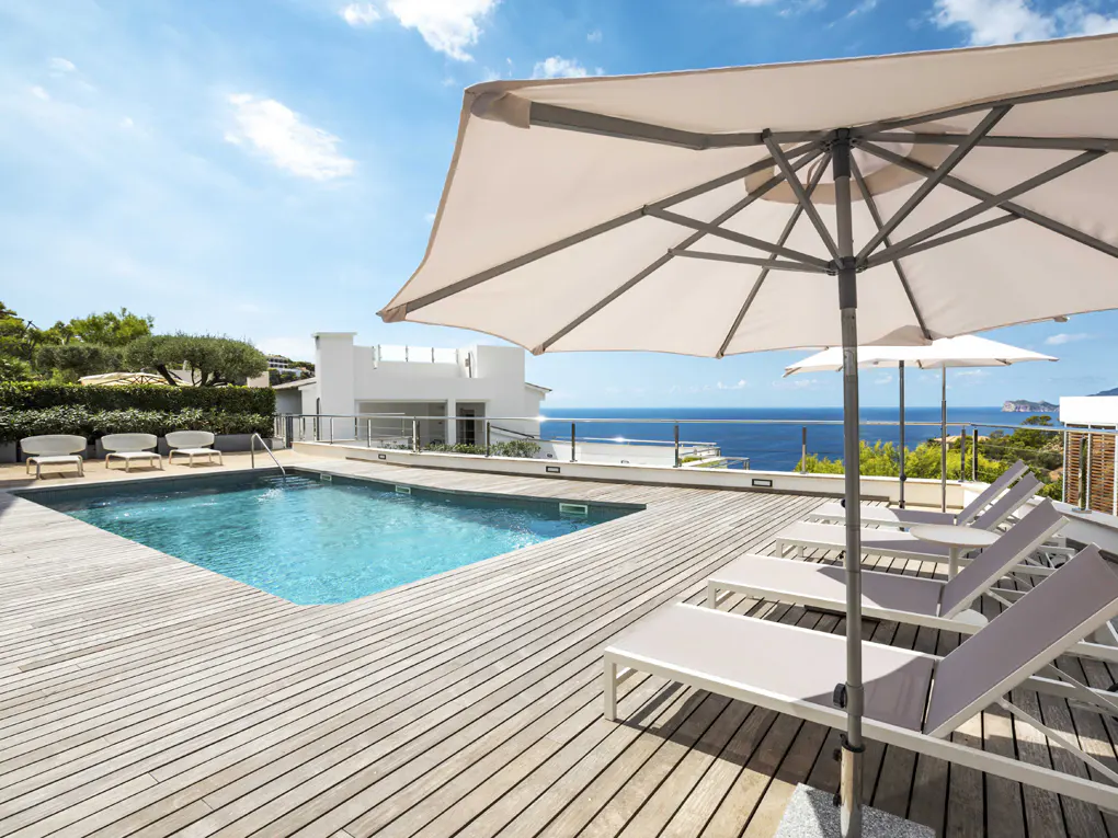 Outdoor pool area with wooden deck, lounge chairs, and umbrellas overlooking the ocean on a sunny day.
