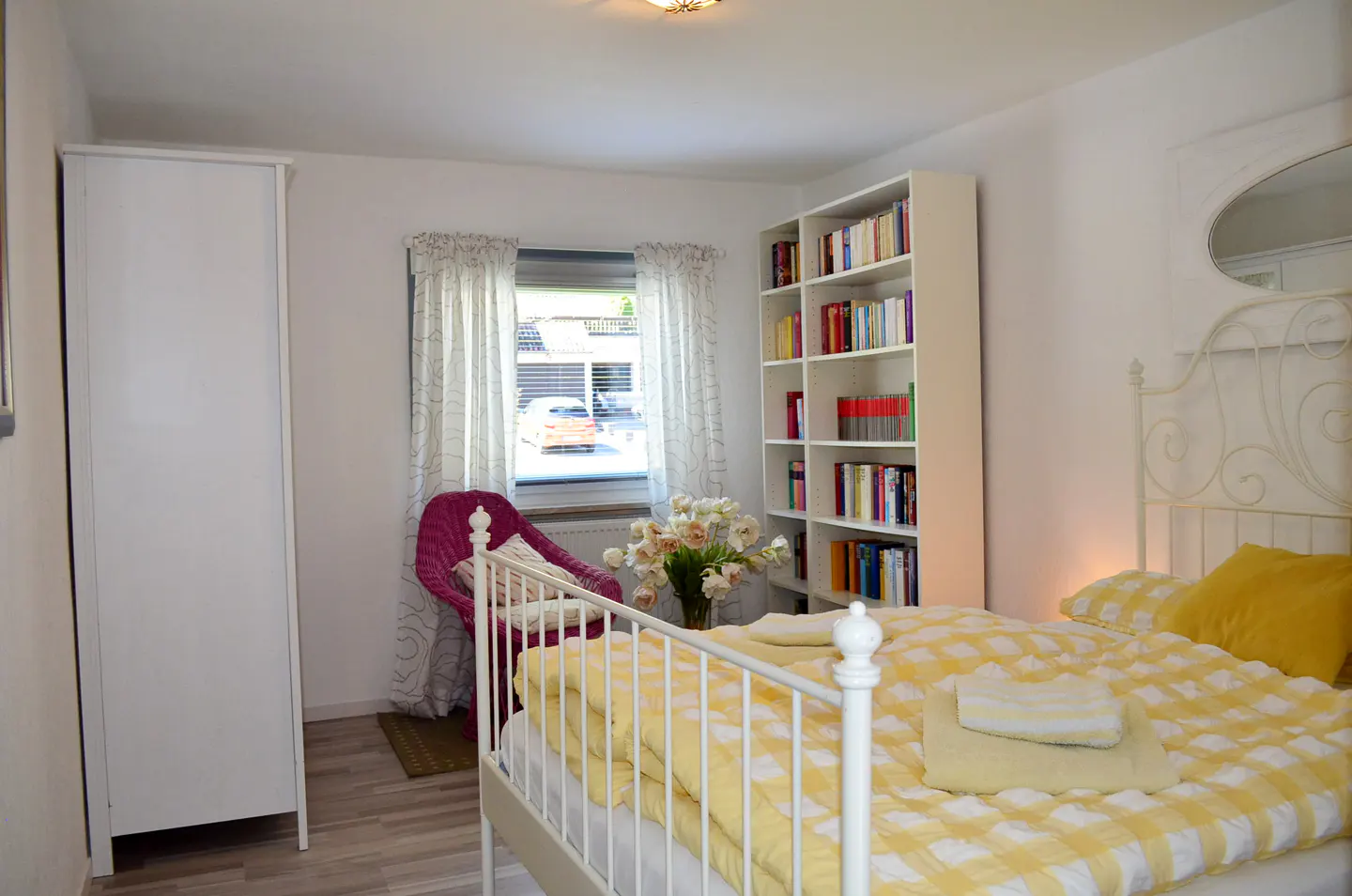 Bright bedroom with a white metal bed, yellow checkered bedding, a tall bookshelf, a pink wicker chair, and a white wardrobe.