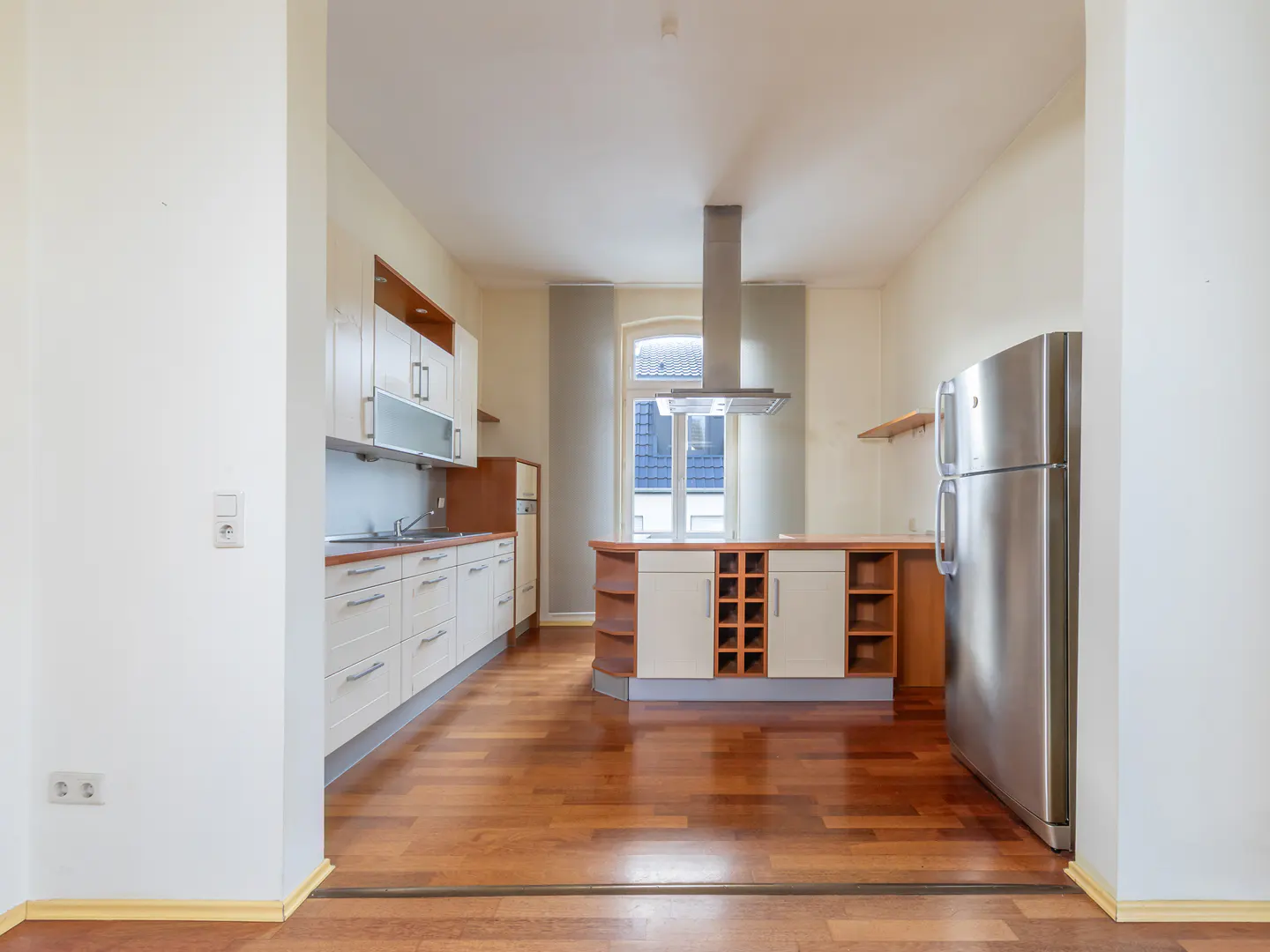 A bright kitchen with white cabinets, a stainless steel refrigerator, and a wooden floor. A kitchen island with a wine rack stands in the center.
