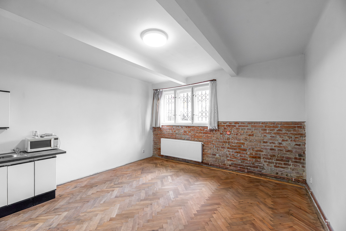 Empty room with herringbone wood floors, white walls, a brick accent wall, a window with security bars, and a kitchenette. [AI-generated text]