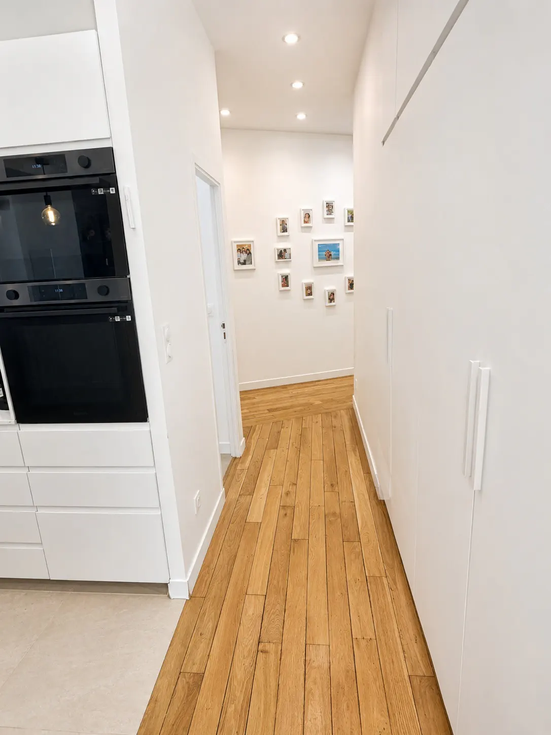 Hallway view with light wood floors, white walls, and recessed lighting. A wall of framed photos is visible at the end of the hall.