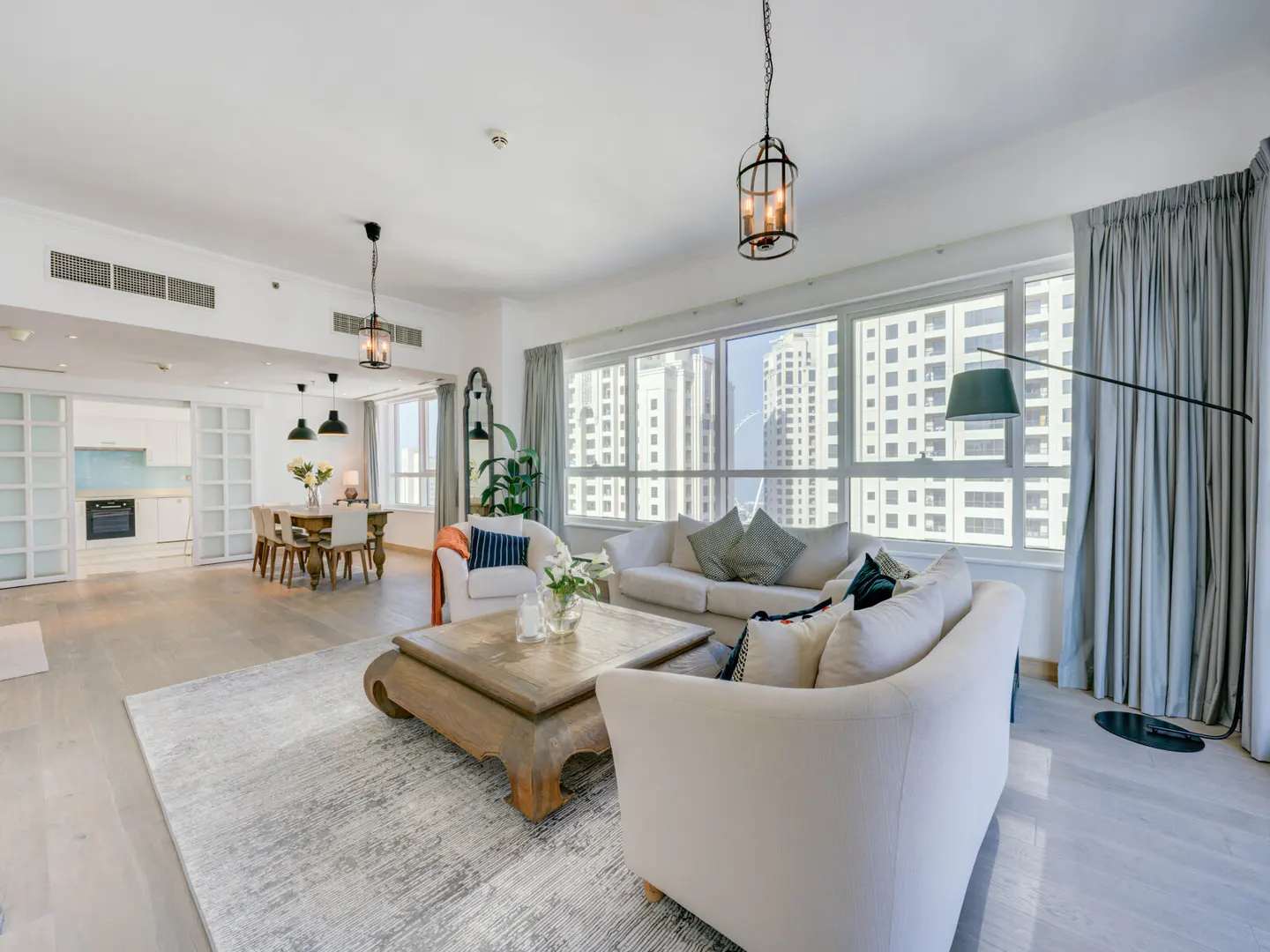 Bright, open-plan living room with white sofas, a wooden coffee table, and a dining area visible in the background. Large windows offer a city view.