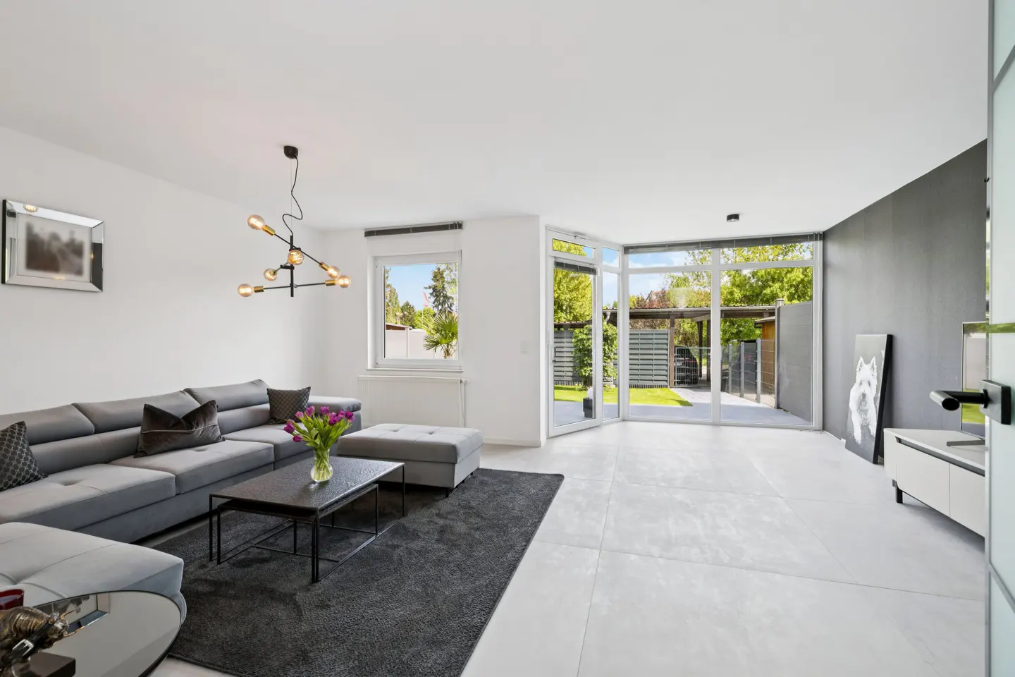 Bright living room with gray sectional sofa, black coffee table with purple flowers, and large windows overlooking a green backyard.