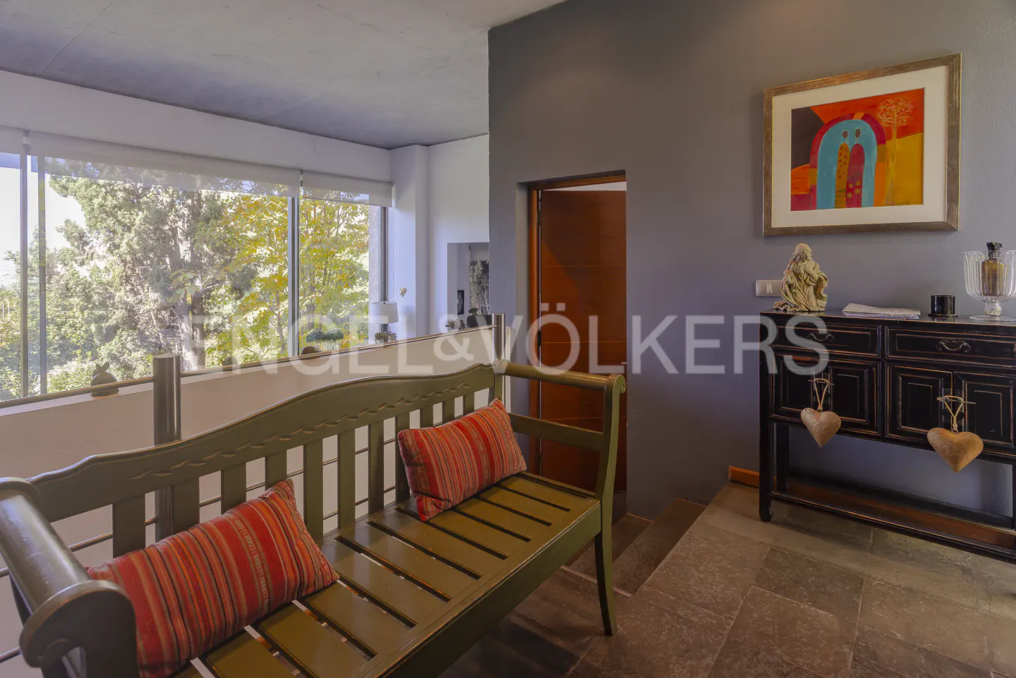 Interior view of a home with a green bench, striped pillows, and a large window with trees outside. A dark wood cabinet and colorful art are on the wall.