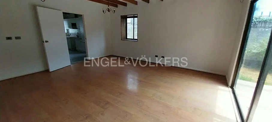 Empty room with wood floors, white walls, and exposed beams. A door leads to a kitchen. A window and glass door provide natural light.
