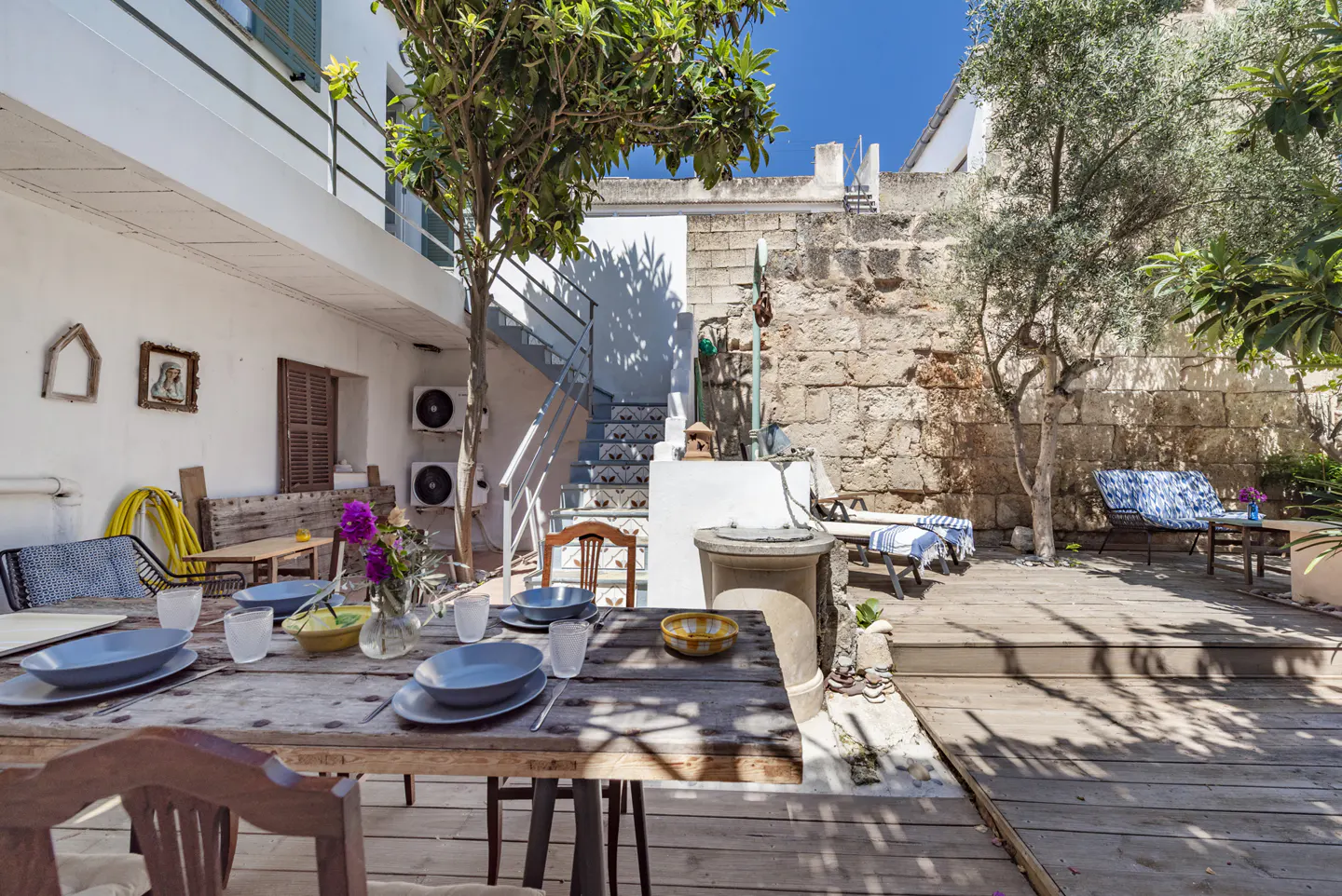 Outdoor dining area with a wooden table set with blue plates, glasses, and flowers. Stone wall, trees, and seating in the background.