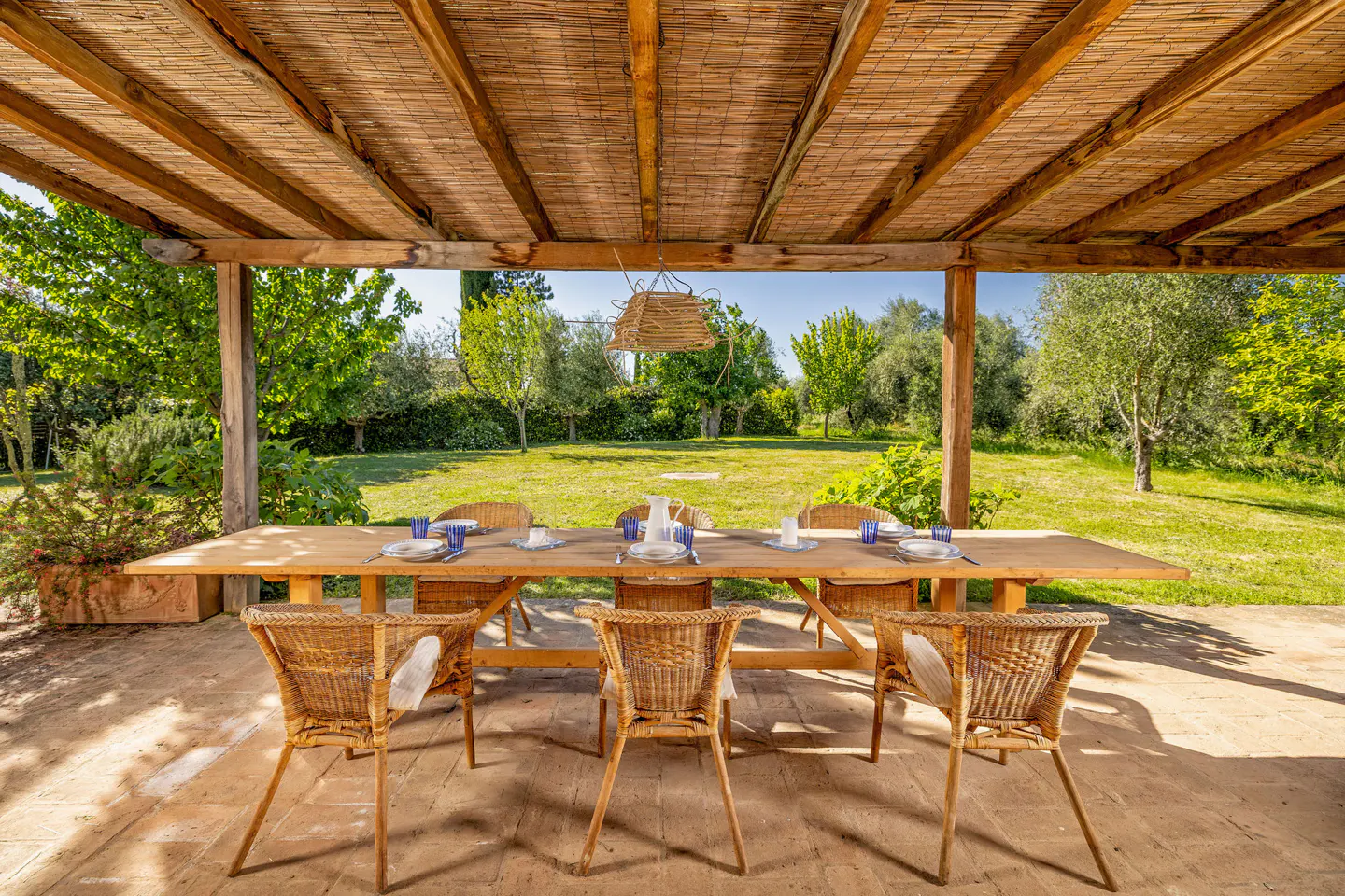 Outdoor dining area with a long wooden table and wicker chairs under a thatched roof, overlooking a green lawn and trees.