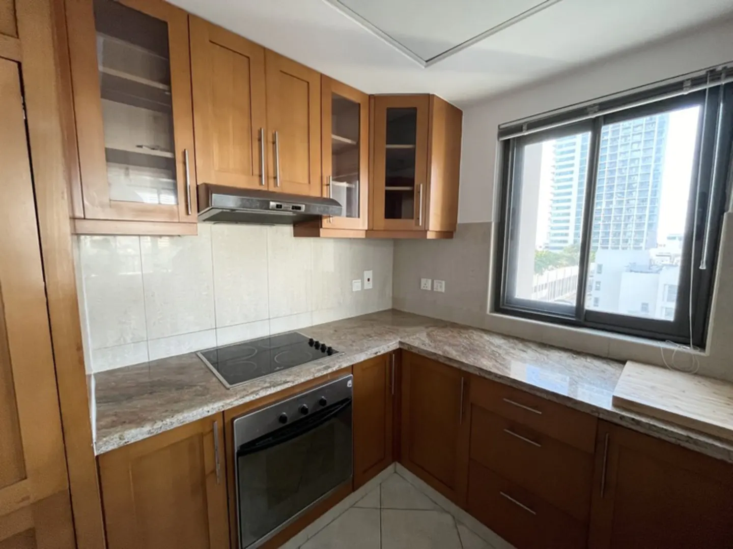 A kitchen with light brown cabinets, granite countertops, and a black stovetop. A window shows a city view.