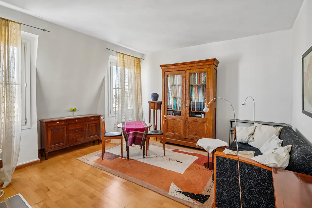 Living room with wooden floors, a patterned rug, a wooden cabinet, a dining set, and a black patterned sofa.