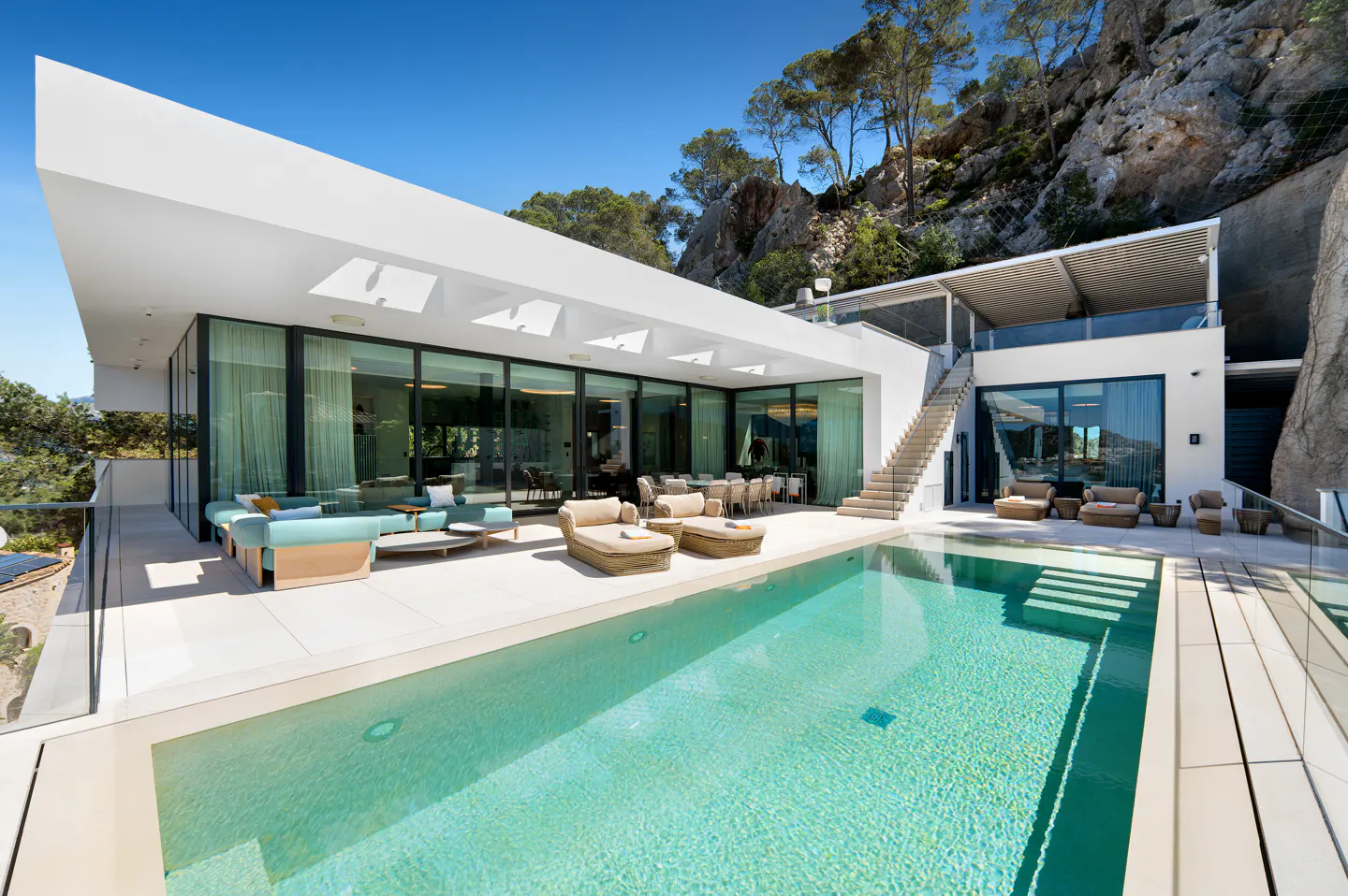 Modern white villa with a turquoise pool, lounge seating, and a dining area. A stone cliff rises in the background.