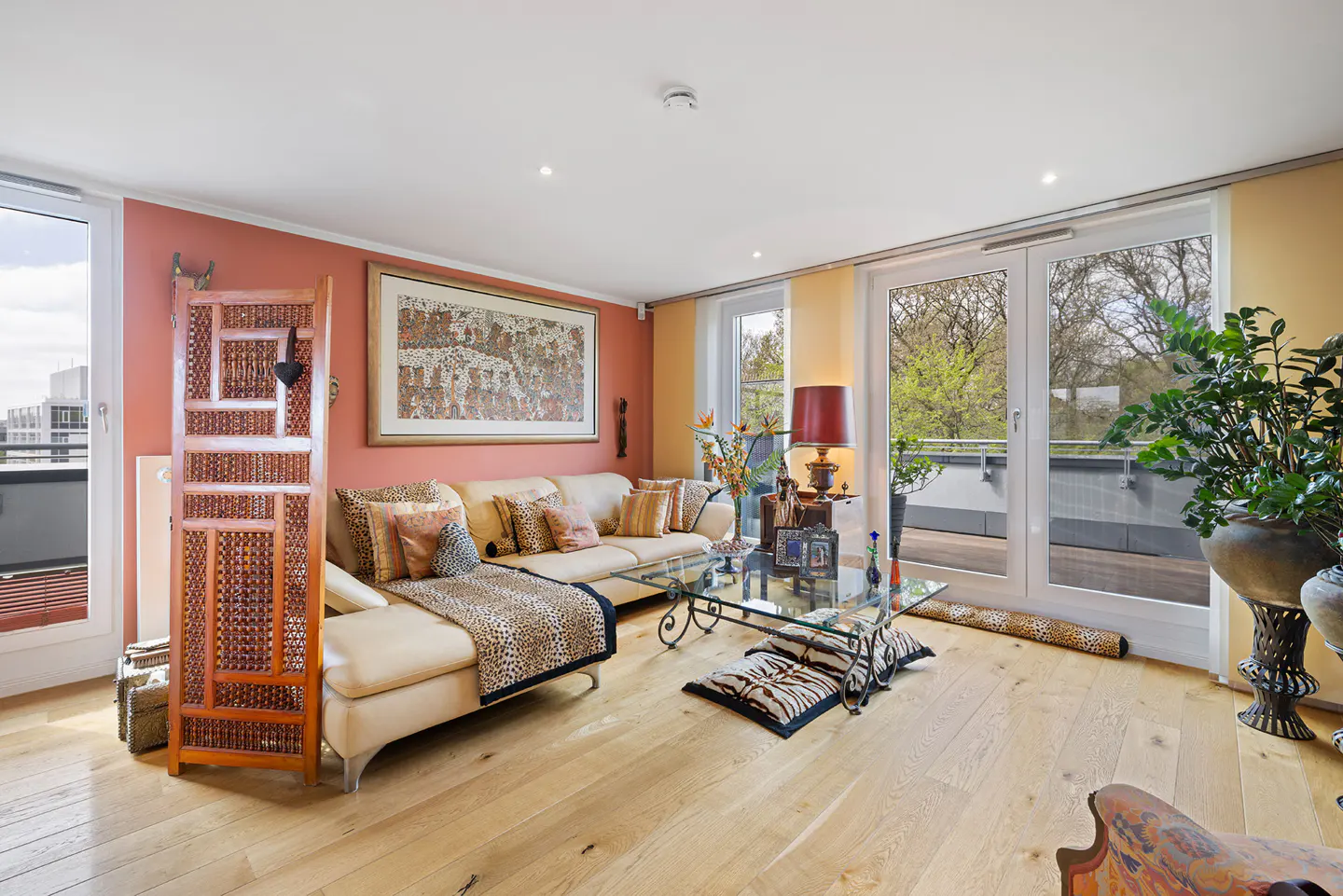Living room with light wood floors, a cream sofa with leopard print throw, and a glass coffee table. Balcony doors let in natural light.