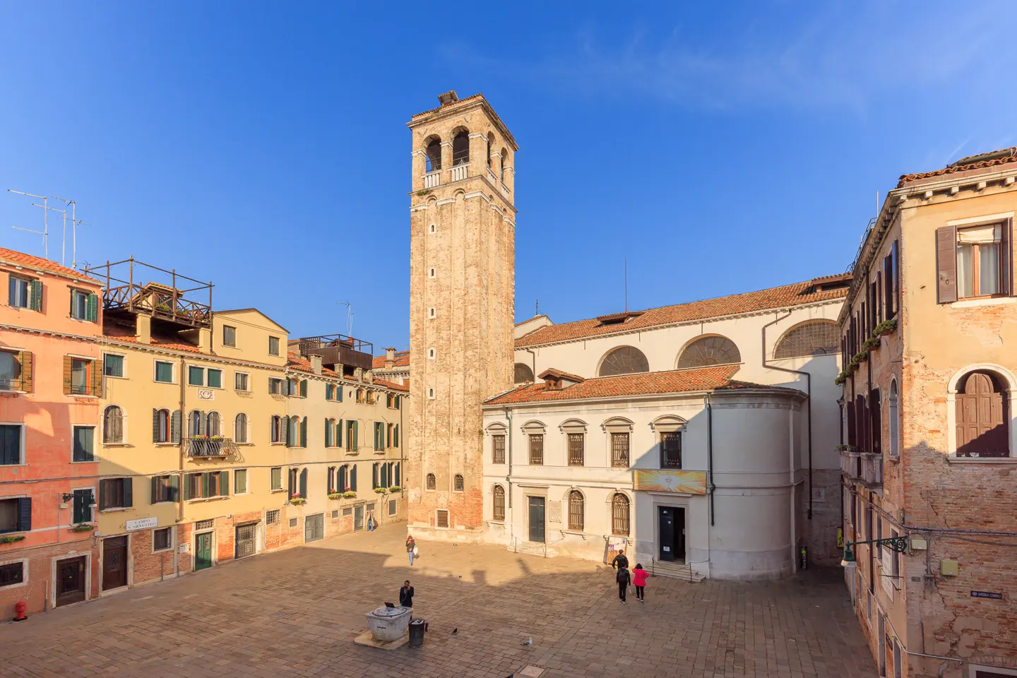 A tall, weathered stone bell tower rises above a sunlit Venetian square surrounded by historic yellow and peach buildings.