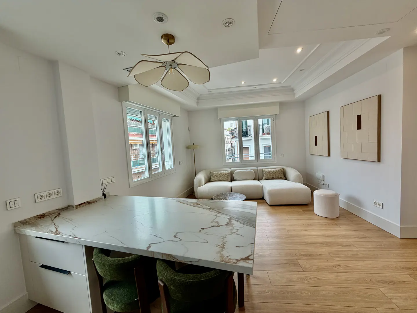 Bright, modern living space with white walls, wood floors, and large windows. A marble countertop island with green chairs is in the foreground.