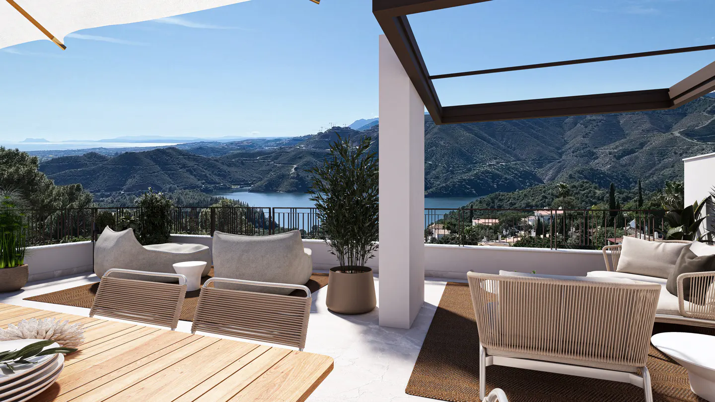 Outdoor patio with mountain view. Wicker chairs and a wooden table sit on a brown rug. A lake and mountains are in the background.