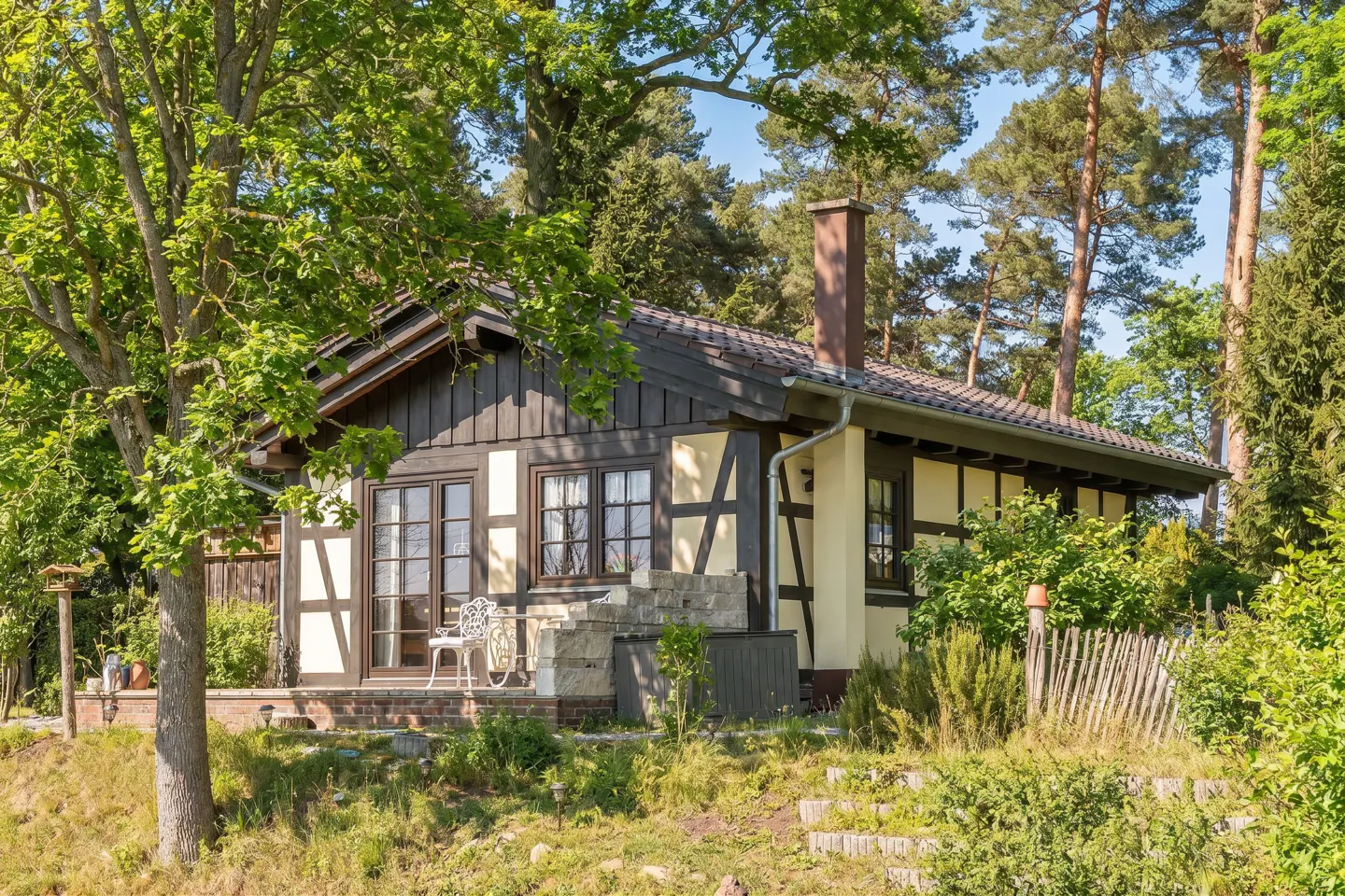 A quaint, half-timbered house with dark wood and cream walls, nestled among lush green trees. A small porch with a white chair is visible.