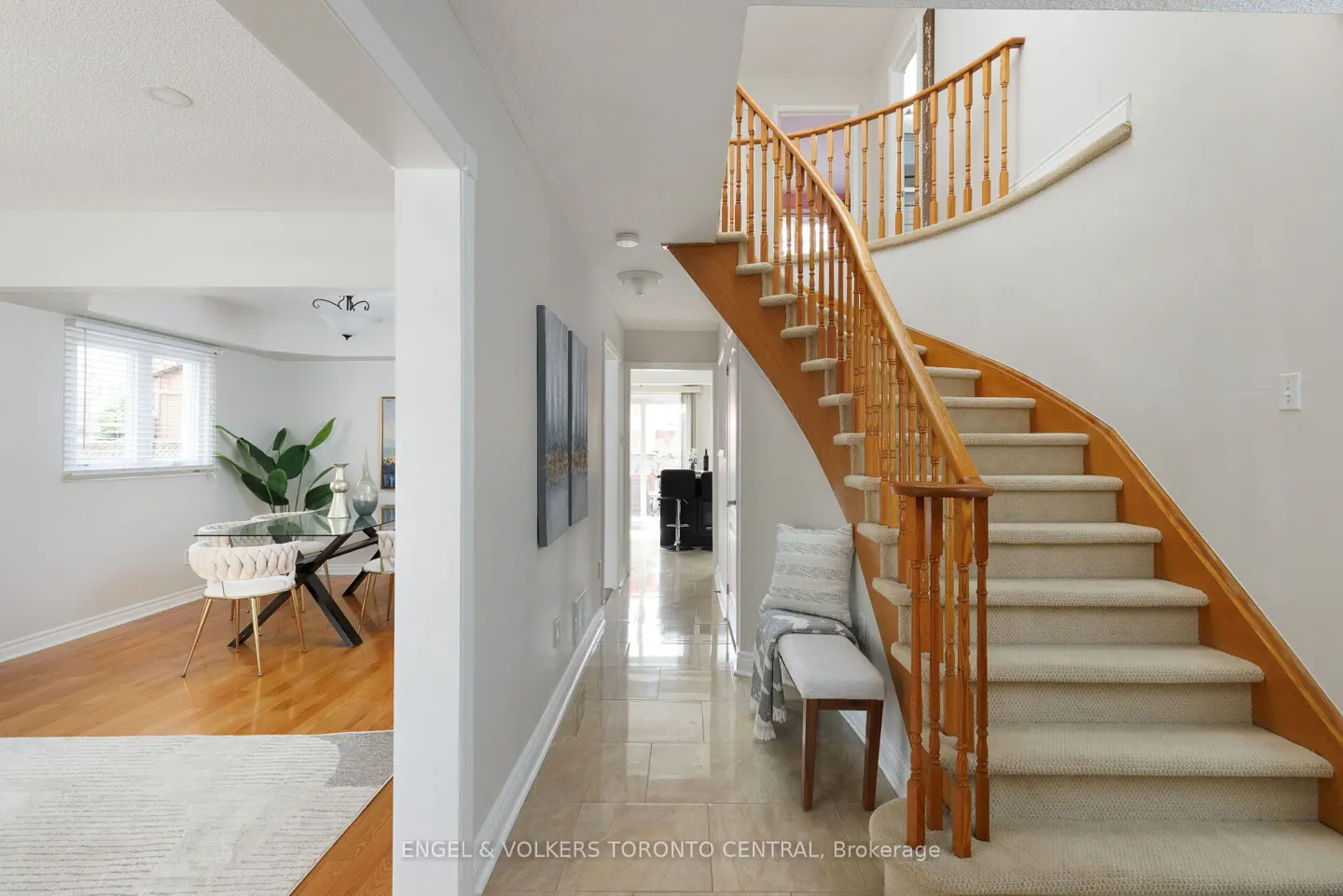 A bright foyer with beige tile floors, a wooden staircase with carpeted steps, and a dining room visible to the left.