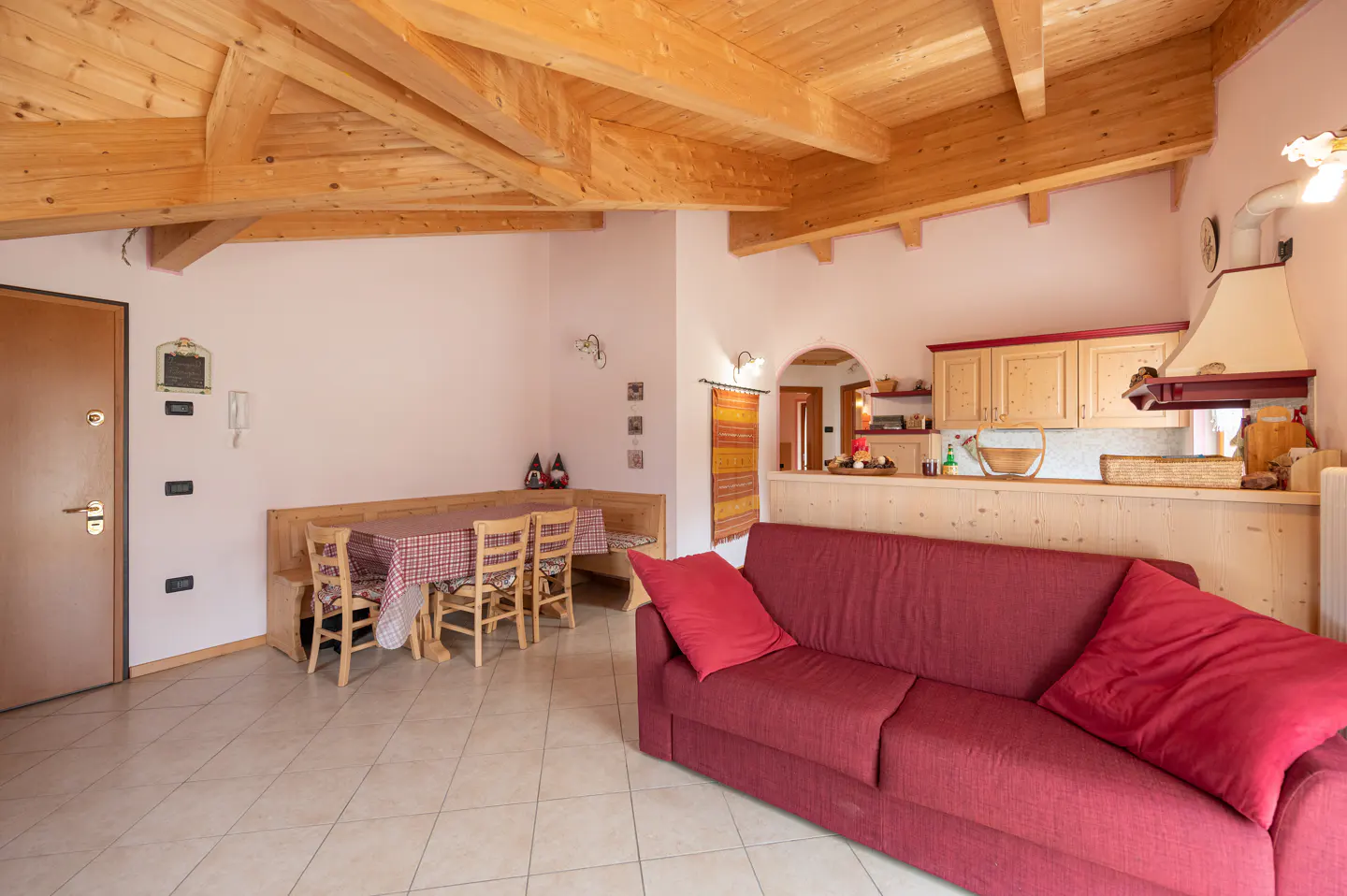Open-concept living space with a red sofa, wooden dining set, and kitchen with light wood cabinets. Exposed wooden beams on the ceiling.