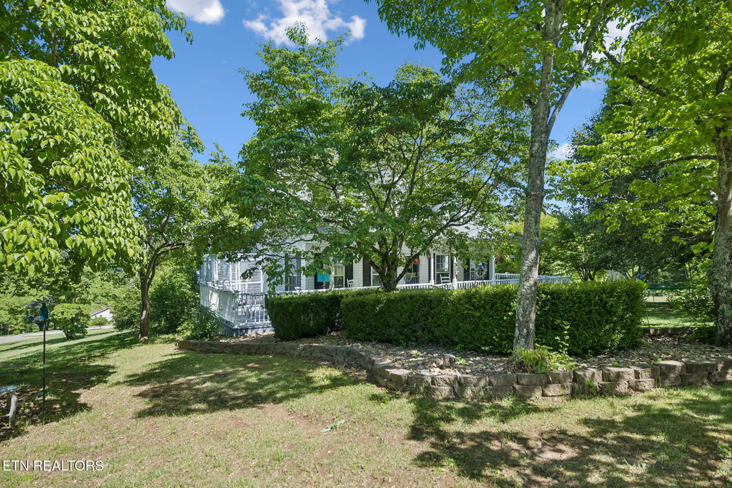 A white house with a porch is partially obscured by green trees and bushes on a sunny day.