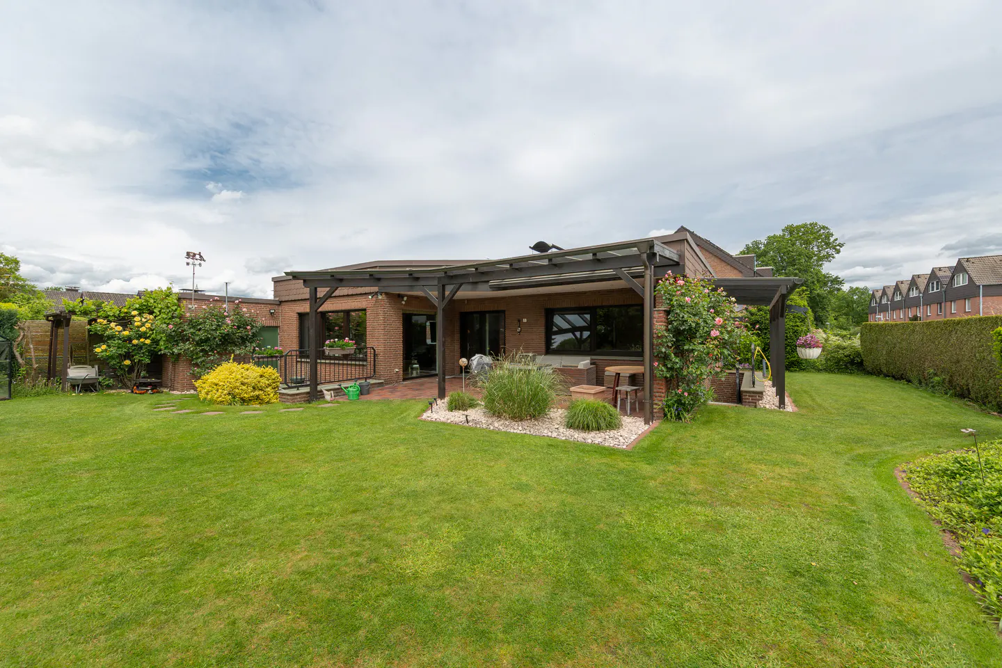 Brick house with a covered patio, lush green lawn, garden beds, and climbing roses under a cloudy sky.