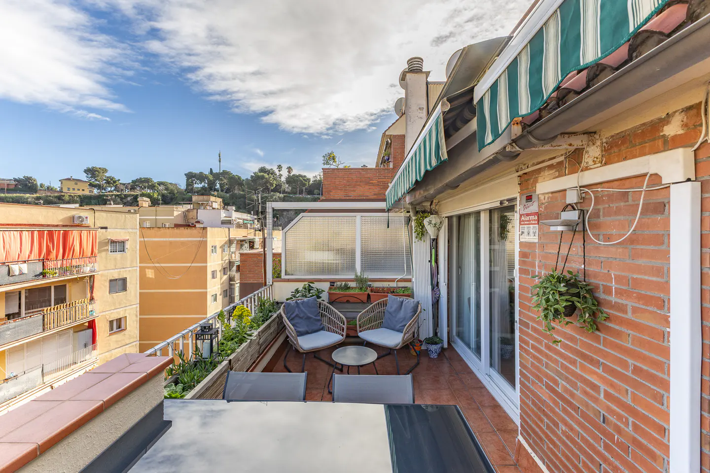 A sunny balcony with two wicker chairs, a small table, and a dining table, overlooking a cityscape.