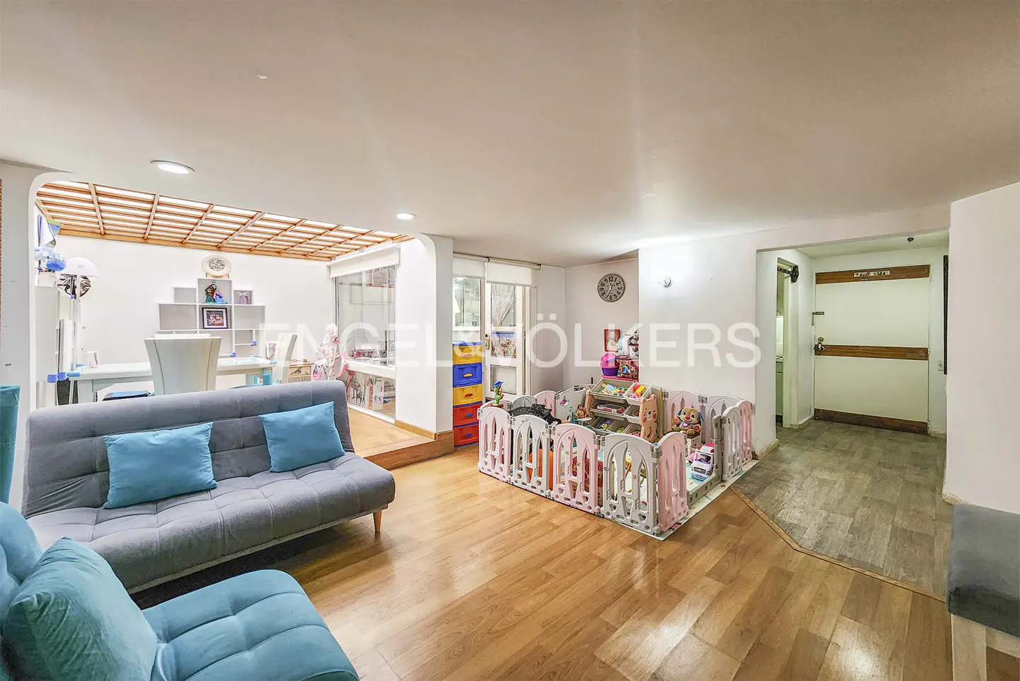 A bright, open living space with hardwood floors, a gray sofa with blue pillows, and a child's play area with a pink and white fence.