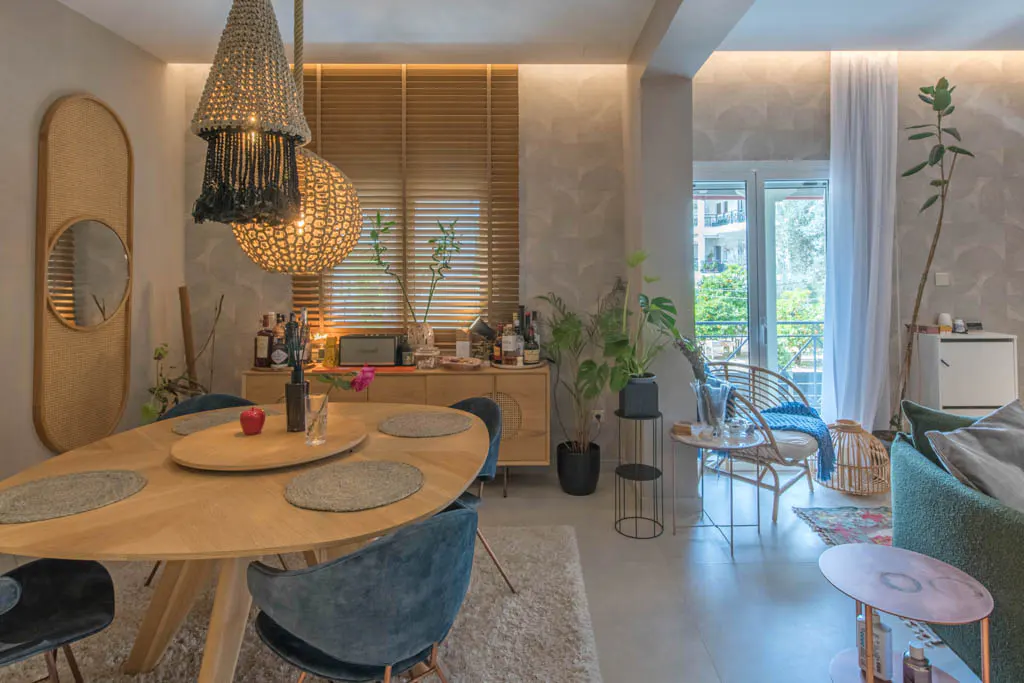 A bright, modern dining and living room with a wooden table, blue chairs, and woven light fixtures. A rattan chair sits near a balcony with a view.