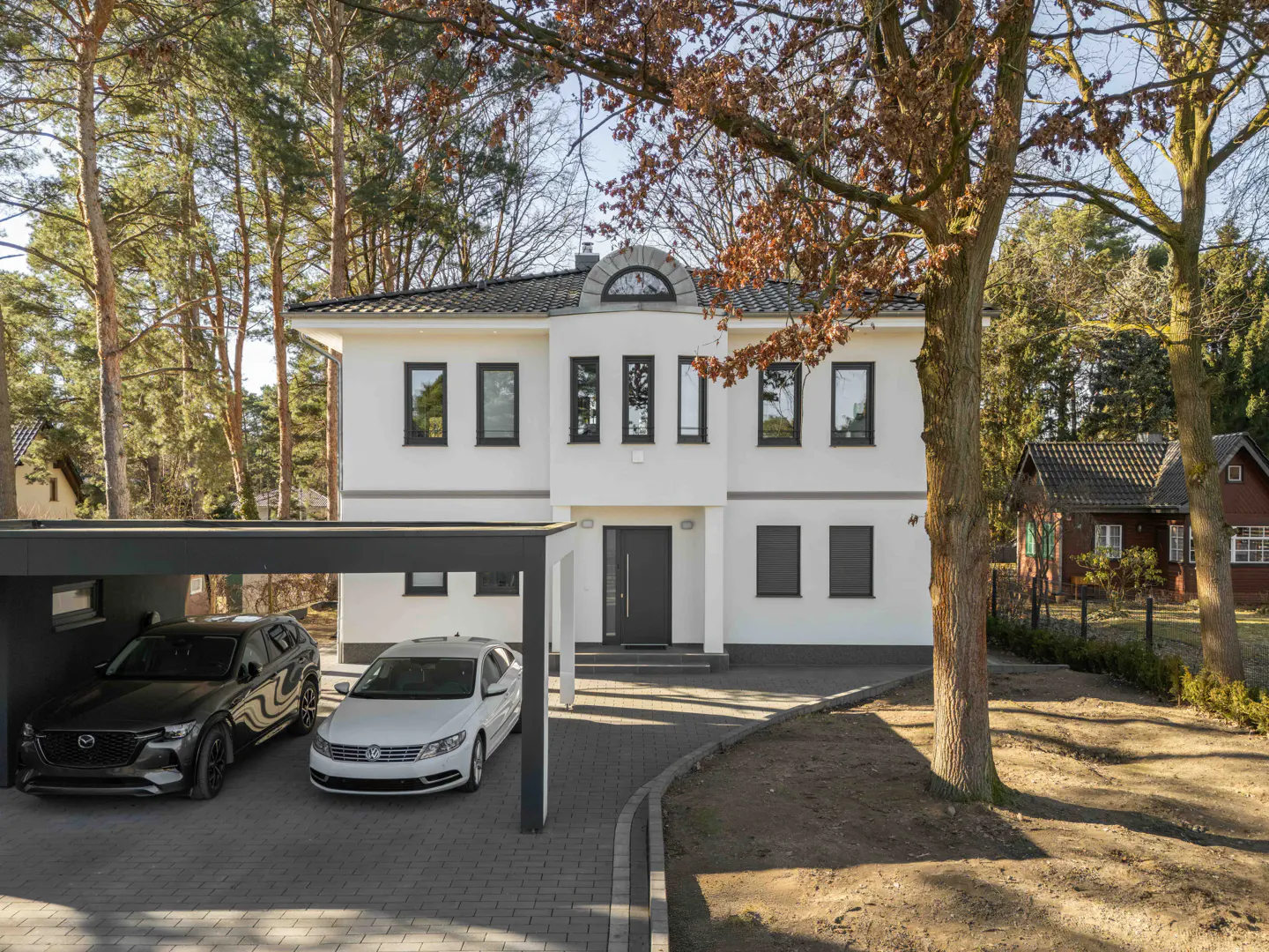 Two-story white house with black trim, a carport with two cars, and a brick driveway. Trees surround the property.