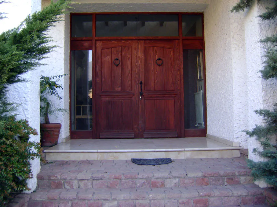 Exterior view of a house entrance with a double wooden door, brick steps, and white stucco walls.