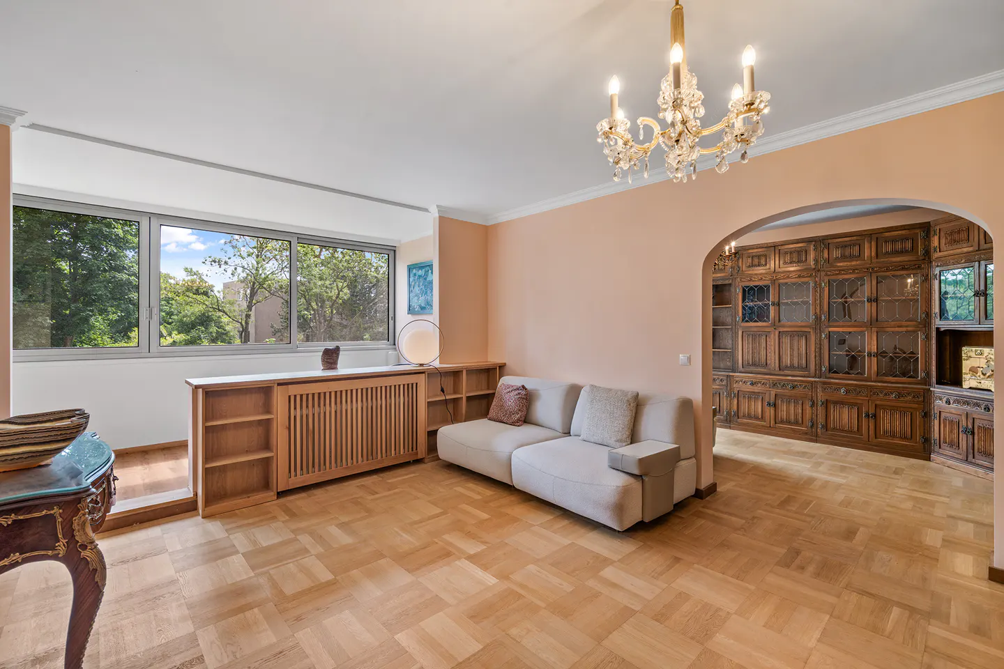 Living room with parquet floors, a beige sofa, and a large window overlooking a green park. A chandelier hangs from the ceiling.