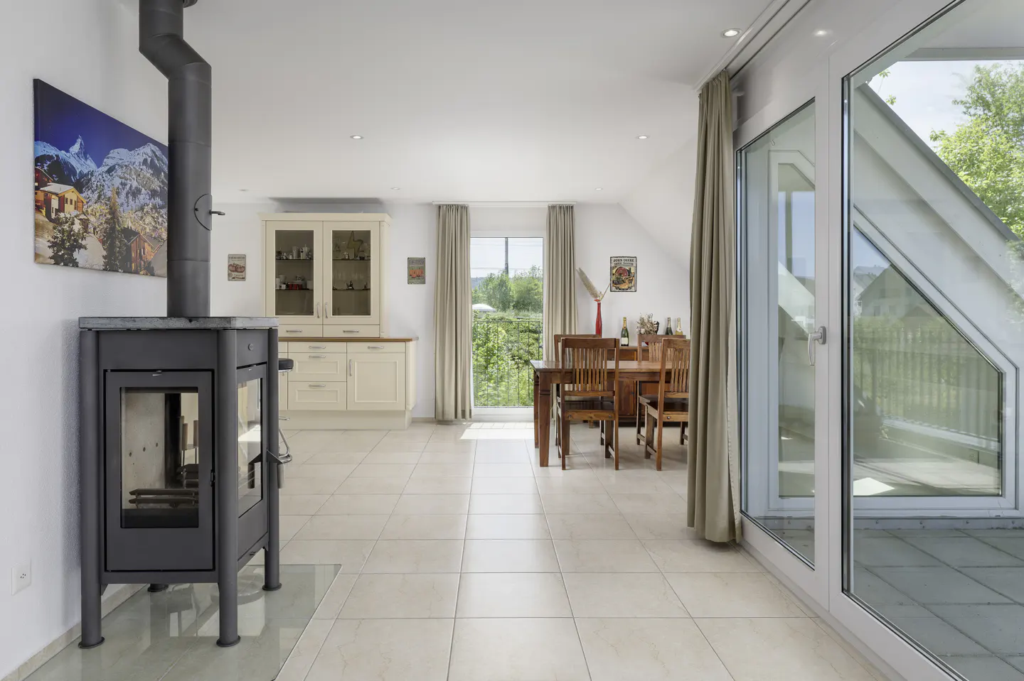 Bright, open-plan living space with a black wood-burning stove, dining table, and cream kitchen cabinets. Large windows offer a view of greenery.