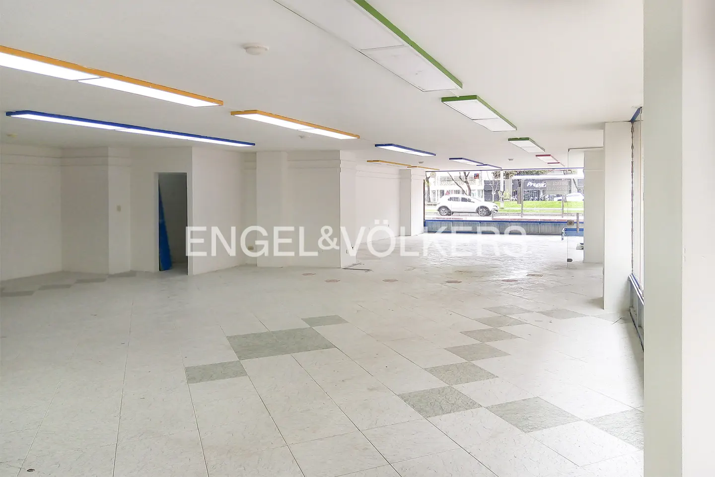 Empty commercial space with white tile floor, white walls, and colorful lights. Engel & Völkers logo in the center.