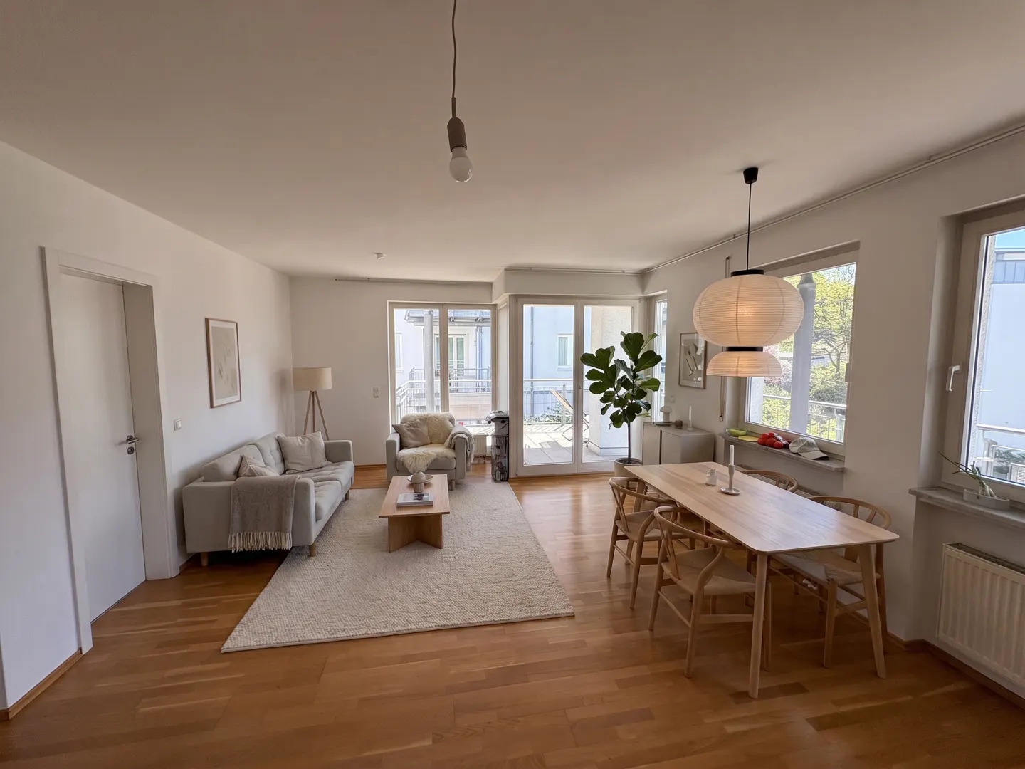 Bright, open-plan living and dining area with wooden floors, a light sofa, a dining table with chairs, and a large window.