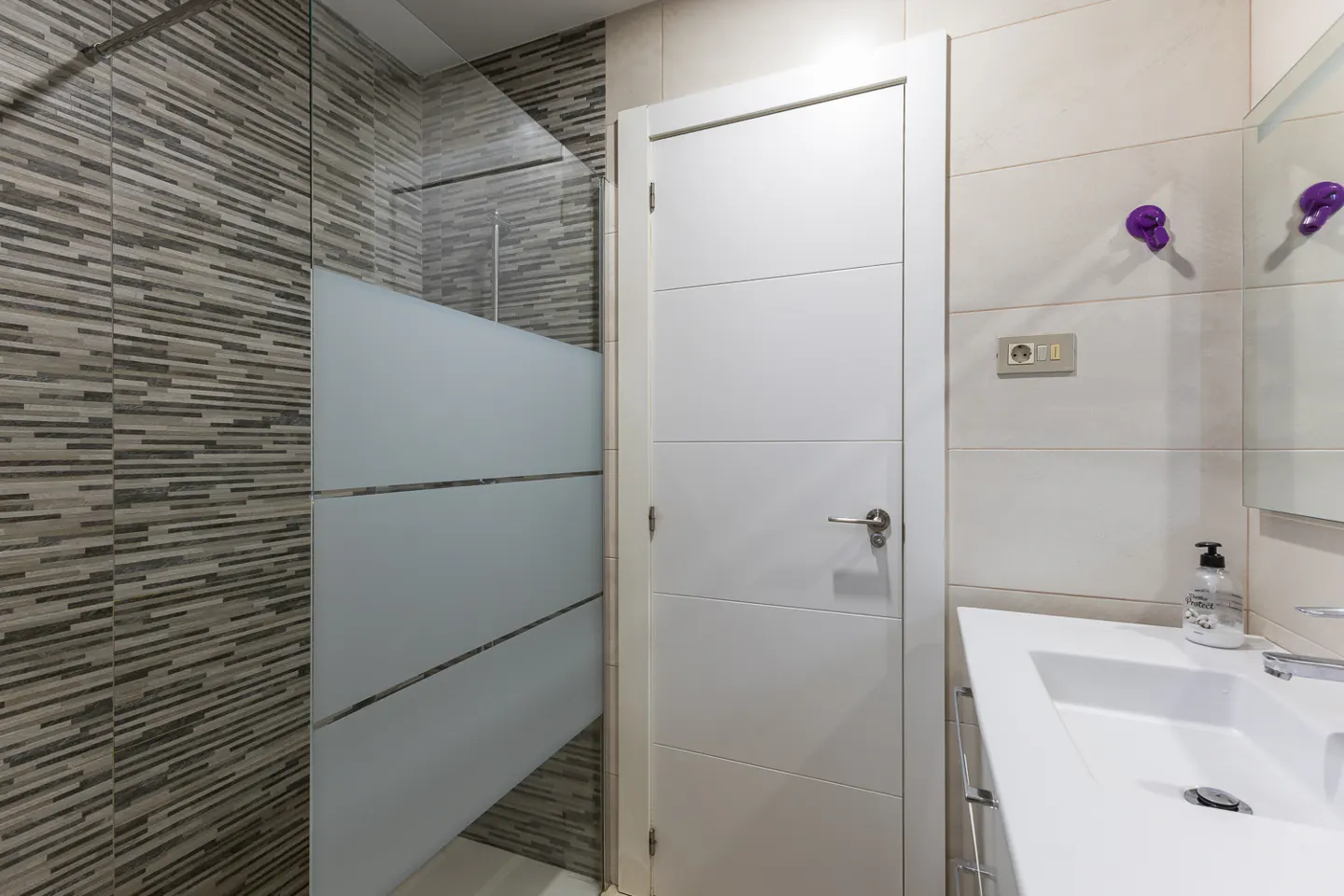 Modern bathroom with a white door, glass shower stall with grey textured tiles, and a white vanity sink.
