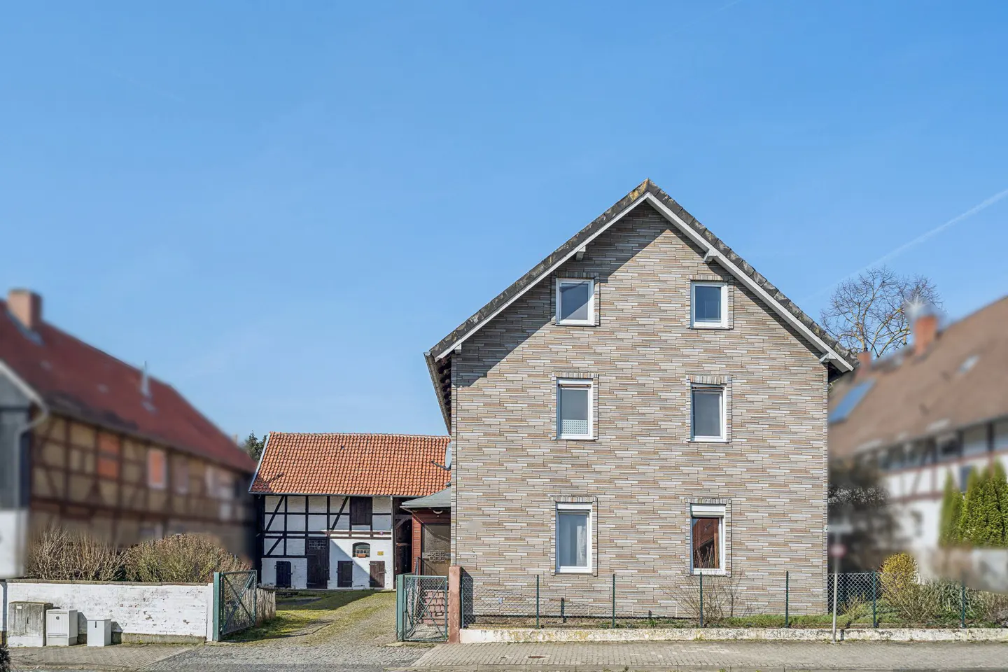 Two-story house with a gray brick facade and white-framed windows, set against a clear blue sky. Other houses are visible in the background.