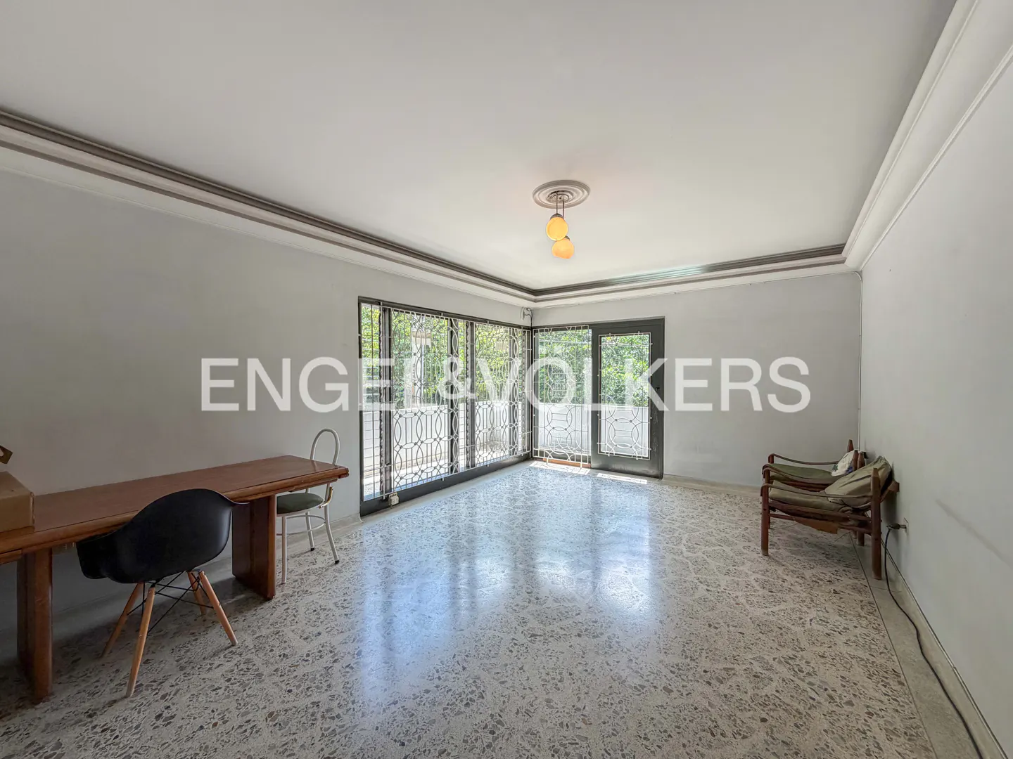 A bright room with a desk, chairs, and a large window with decorative bars. The floor is tiled with a speckled pattern.