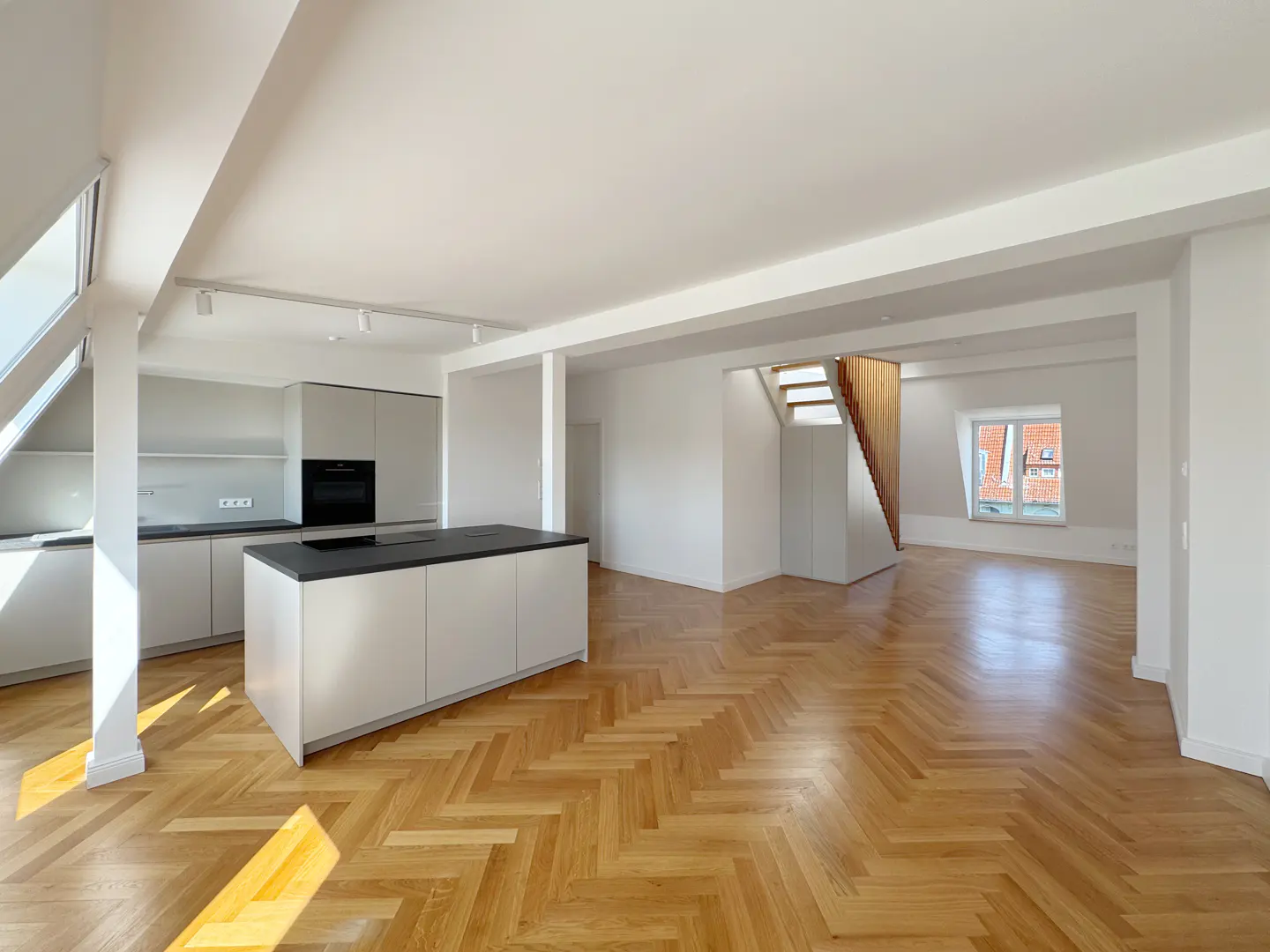 Bright, open-concept apartment with herringbone wood floors, white walls, and a modern kitchen with a black island. Stairs lead to another level.