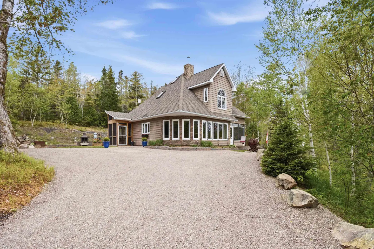 A two-story brown house with a stone foundation and gravel driveway, surrounded by lush green trees under a blue sky.
