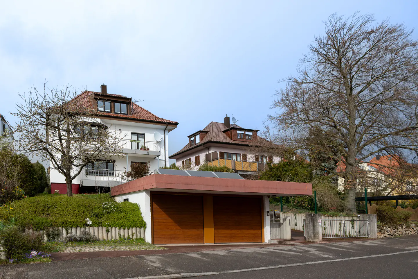Two-story houses with red-tiled roofs sit behind a two-car garage with brown doors and a flat roof. Bare trees frame the scene.