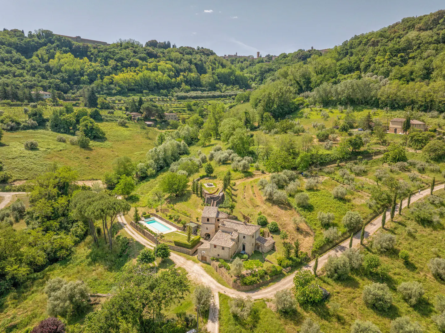 Aerial view of a stone villa with a swimming pool nestled in a lush, green Italian hillside landscape with olive trees.