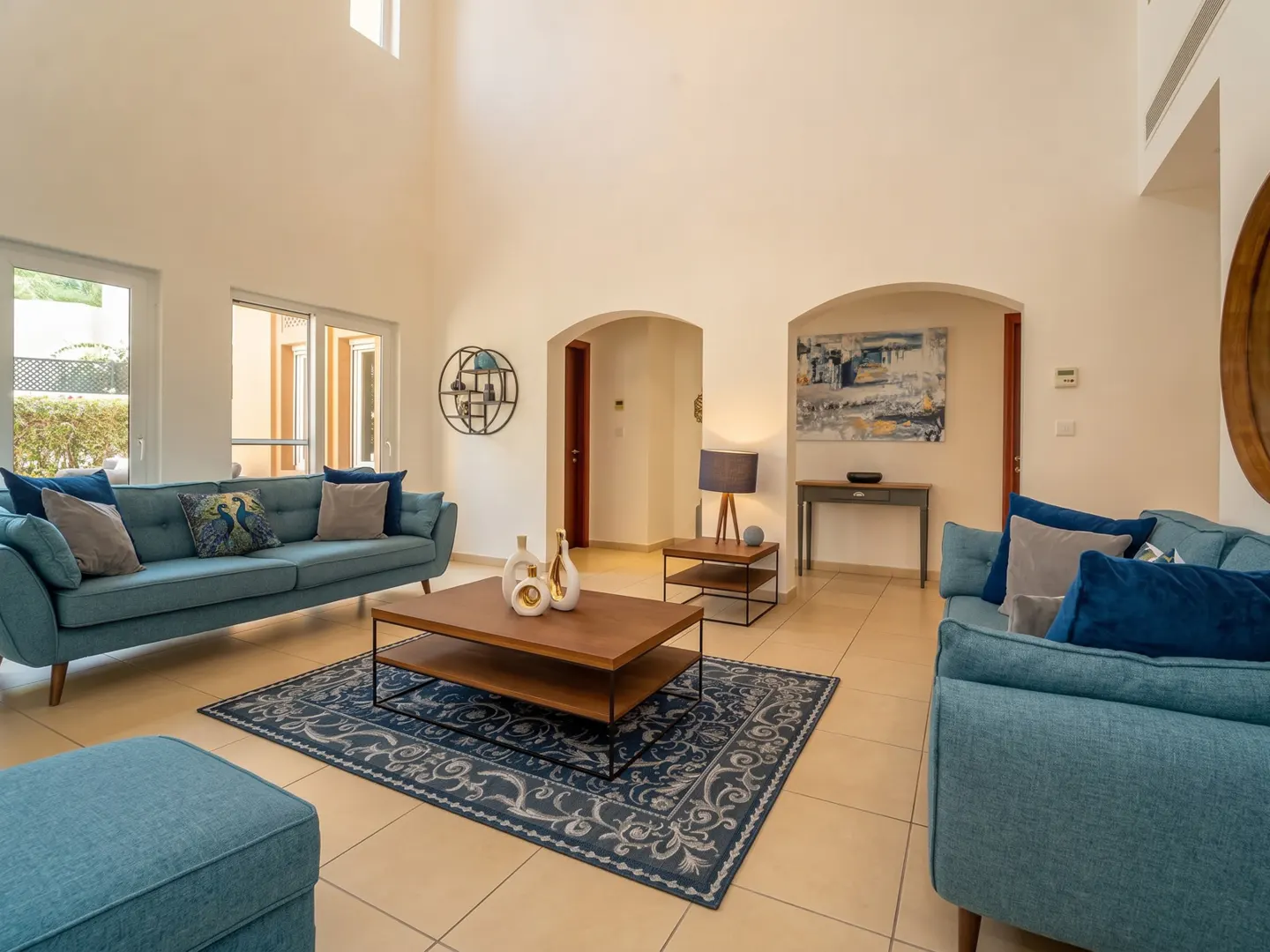 Spacious living room with high ceilings, teal sofas, a wooden coffee table, and a patterned rug on tiled floors.