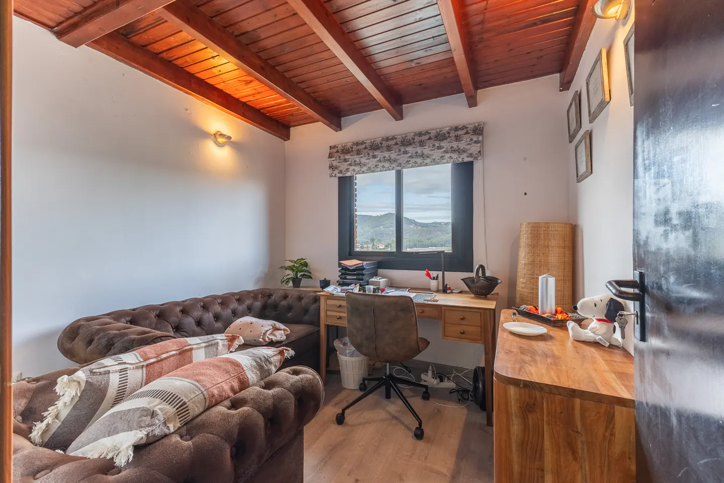 A home office with a brown leather sofa, wooden desk, and a window with a view of mountains.
