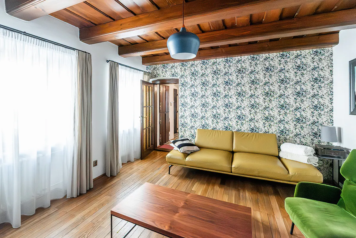 Living room with wood floors, exposed wood beams, floral wallpaper, yellow sofa, green chair, and blue pendant light.